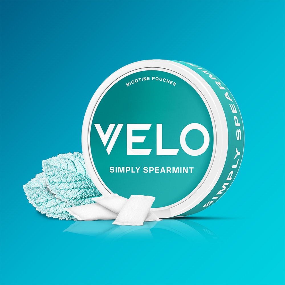 A round container of VELO Simply Spearmint nicotine pouches with three white pouches placed in front of it next to textured mint leaves.