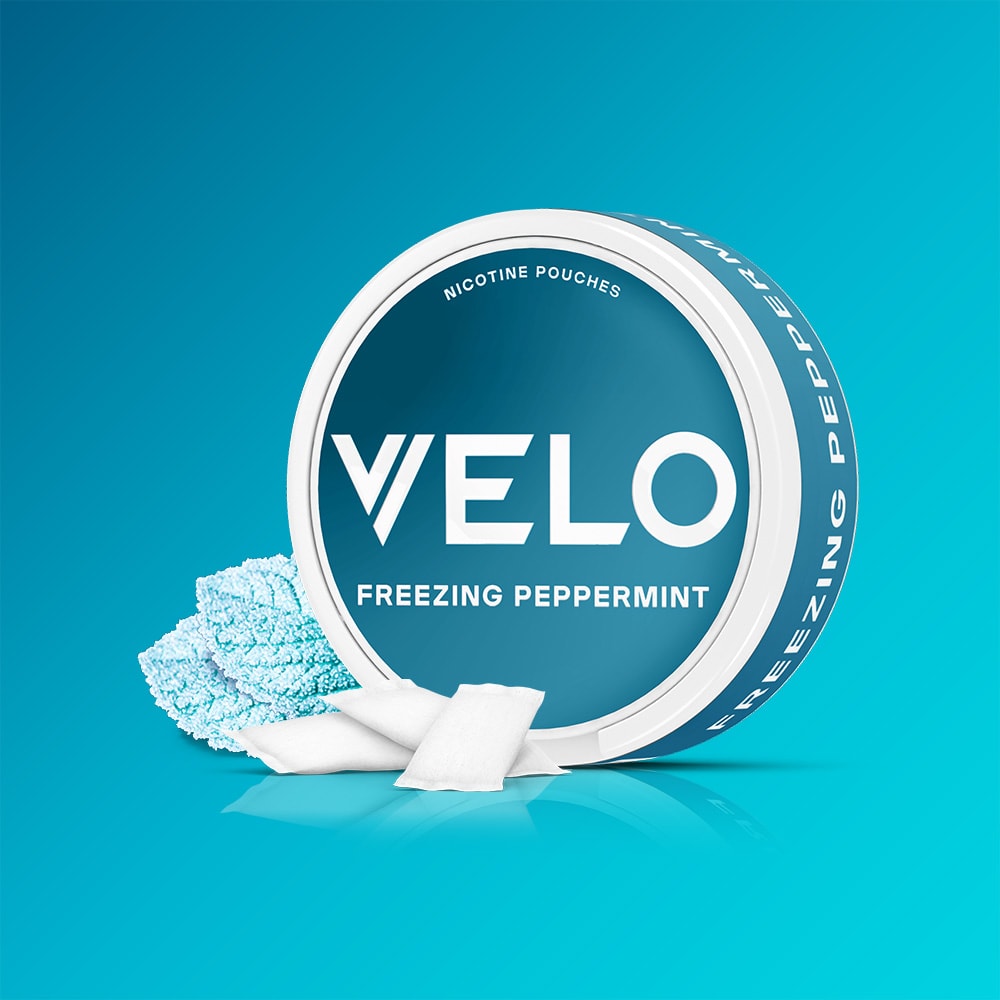 A round container of VELO Freezing Peppermint nicotine pouches next to three white pouches on a blue gradient background.