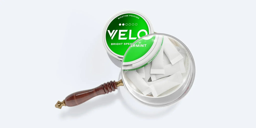 Velo Nicotine Pouches |  A tin of VELO Bright Spearmint nicotine pouches sits open, showing white pouches inside. A magnifying glass is placed over the tin, enlarging part of the Velo pouches and their crisp contents.